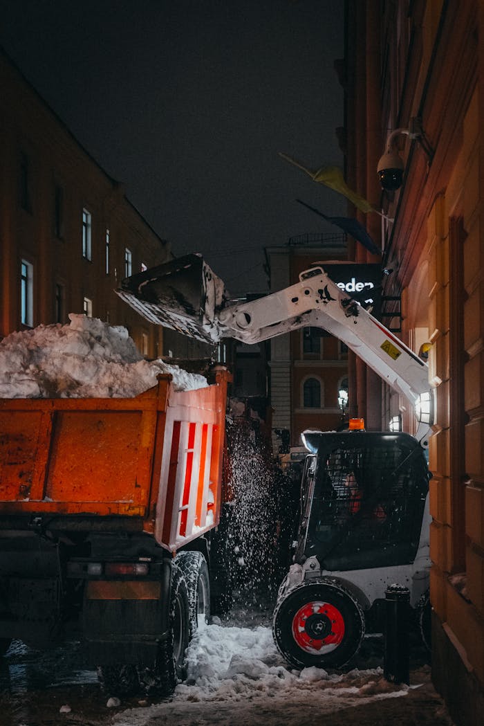 Snow removal in city streets at night, featuring a loader and truck during winter.